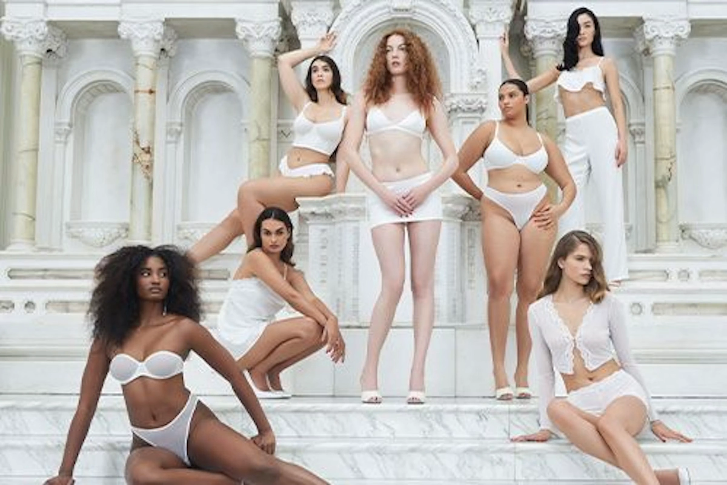 A group of brides and friends in white lingerie and loungewear sit among marble columns